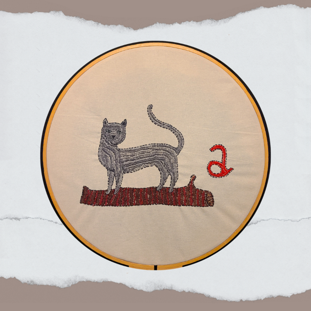 An embroidery project shows a blue-gray cat with green eyes. The cat is standing on a log next to the letter “a” stitched in red. Read together, the cat plus the letter “a” and the log create the title of the piece, Cat-a-log!